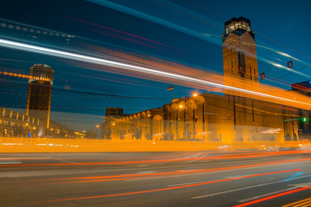 Time lapse photo of traffic at Yerevan city that features blurred lines across a street created from moving vehicles. Featured in the background are some large, historic looking buildings.