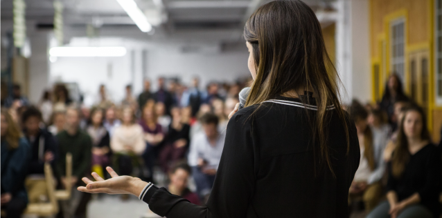 Photo of a woman giving a presentation to a room full of people.
